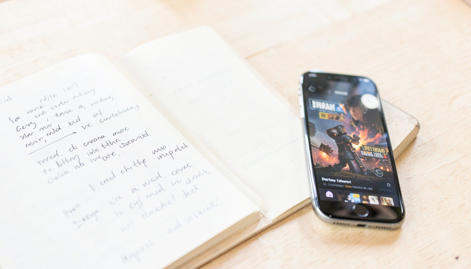 A bright, airy photograph of an open notebook with handwritten notes beside a smartphone showing a game trailer, on a light wooden surface.