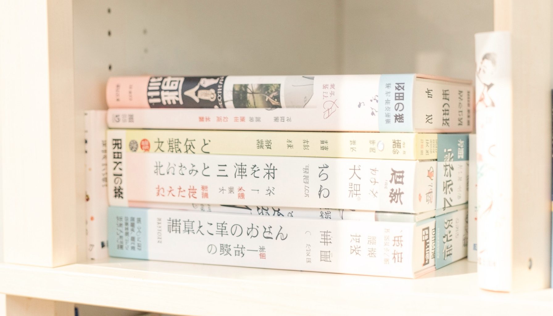 A bright, airy photograph of several Japanese fiction books stacked on a shelf, with soft shadows and warm daylight illuminating their spines.