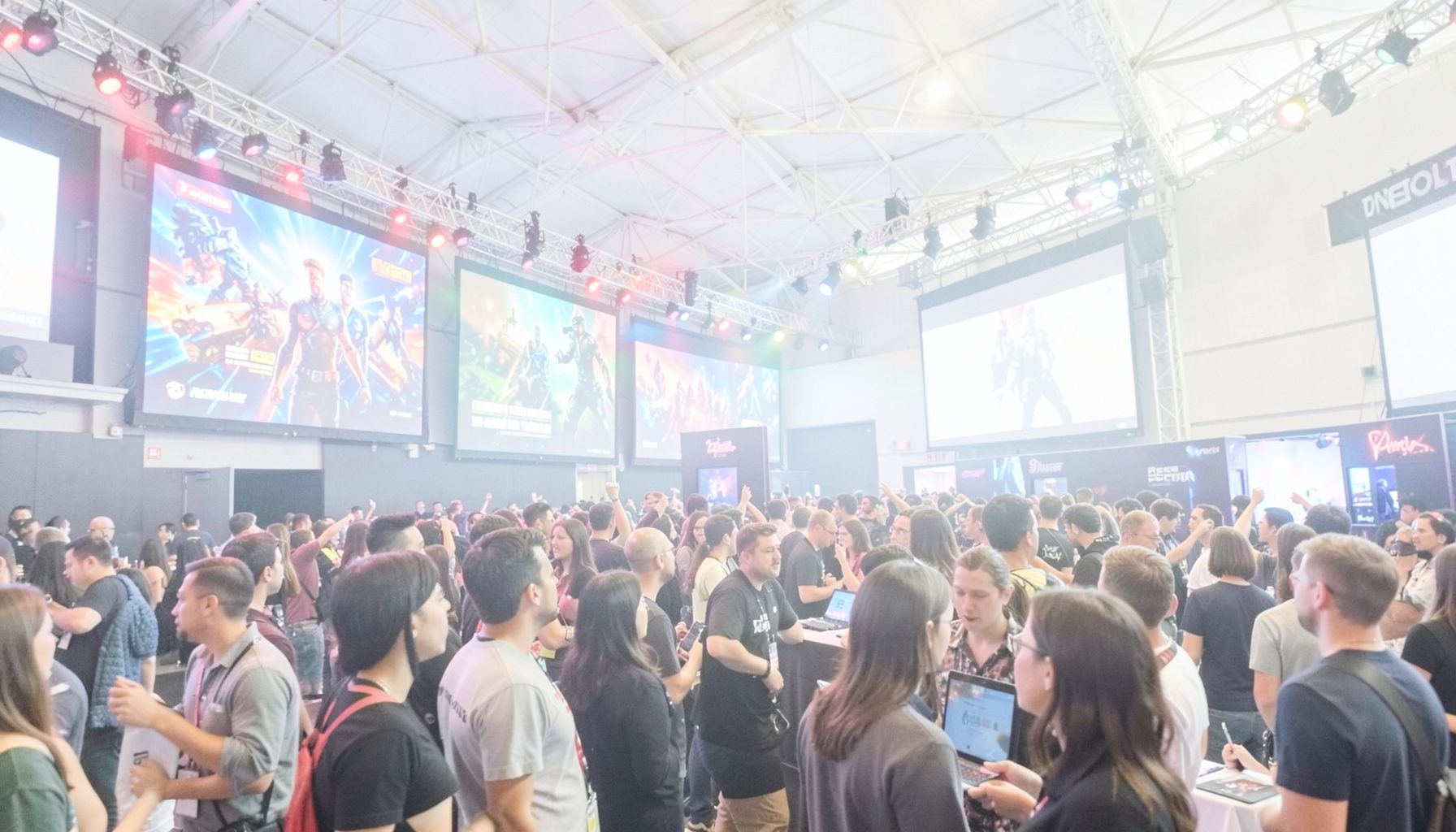 A bright, airy photograph of a gaming convention hall with large screens displaying game trailers, colorful lights, and an excited crowd atmosphere.