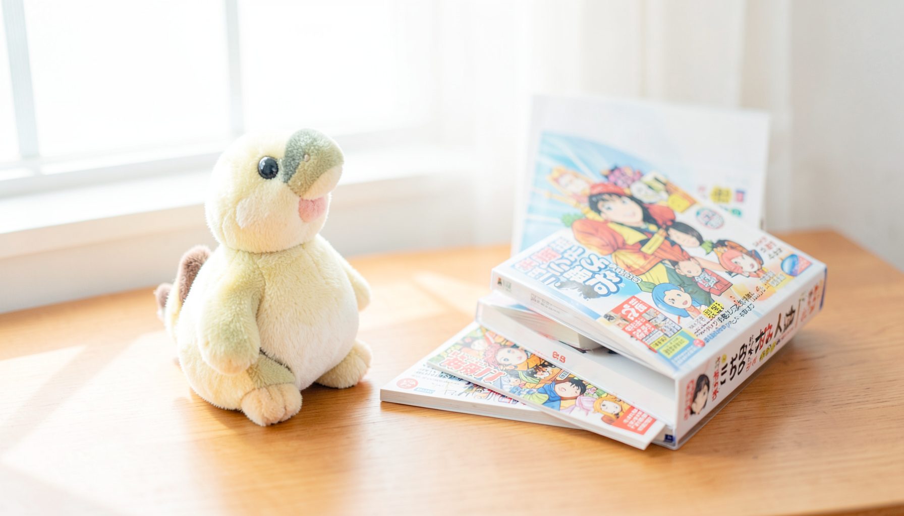 A bright, airy photograph of a tidy desk with Japanese pop-culture collectibles — a small plush chocobo, a manga volume, and a game case — arranged on a warm wooden surface with soft natural light from a window.